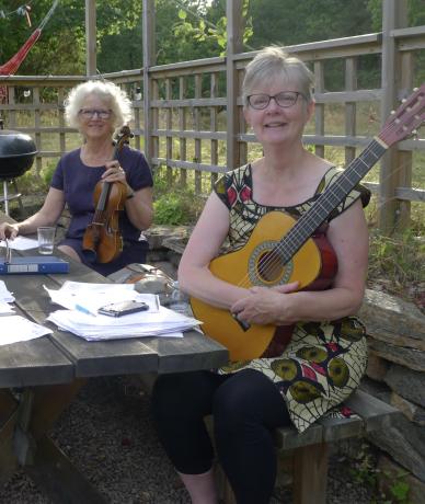 Två kvinnor sitter utomhus vid ett bord, den ena håller i sin fiol och den andra sin gitarr