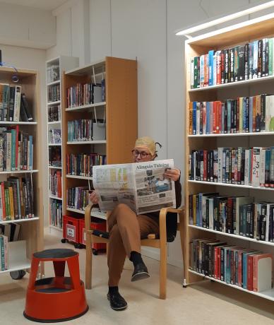 Interiörbild Ingareds bibliotek med hyllor och i en stol sitter en person och läser alingsås tidning
