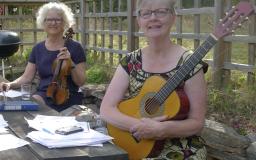 Två kvinnor sitter utomhus vid ett bord, den ena håller i sin fiol och den andra sin gitarr